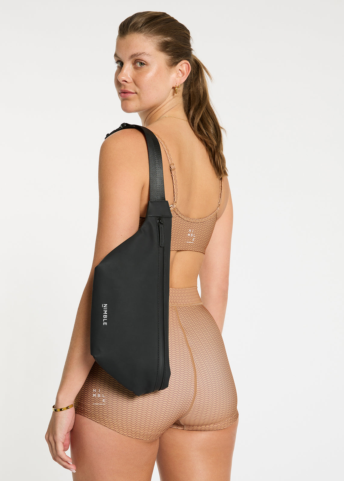 Close Up Of Model Stood Facing Backwards Wearing A Black Splash Proof Cross Body Bag With Contrasting White Nimble Logo To The Centre Bottom Over Their Shoulder.