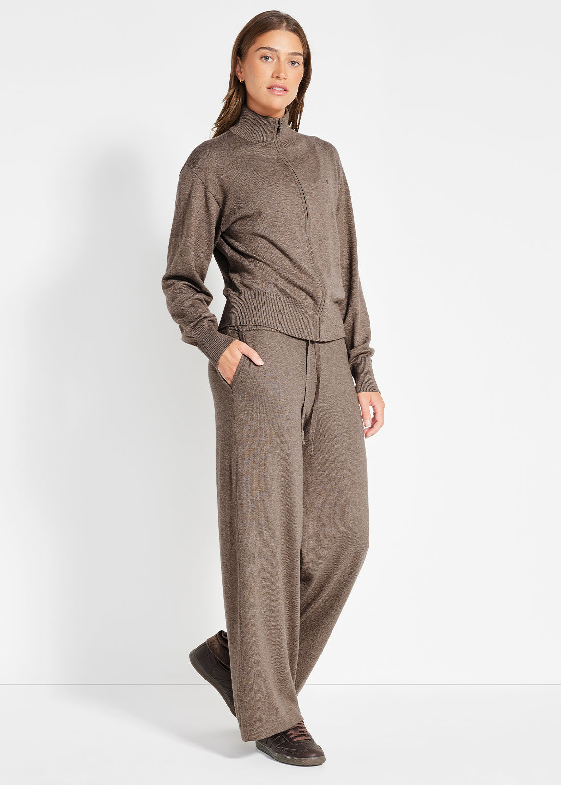 Model wearing camel knit half-zip jumper and matching drawstring pants, barefoot on a sofa