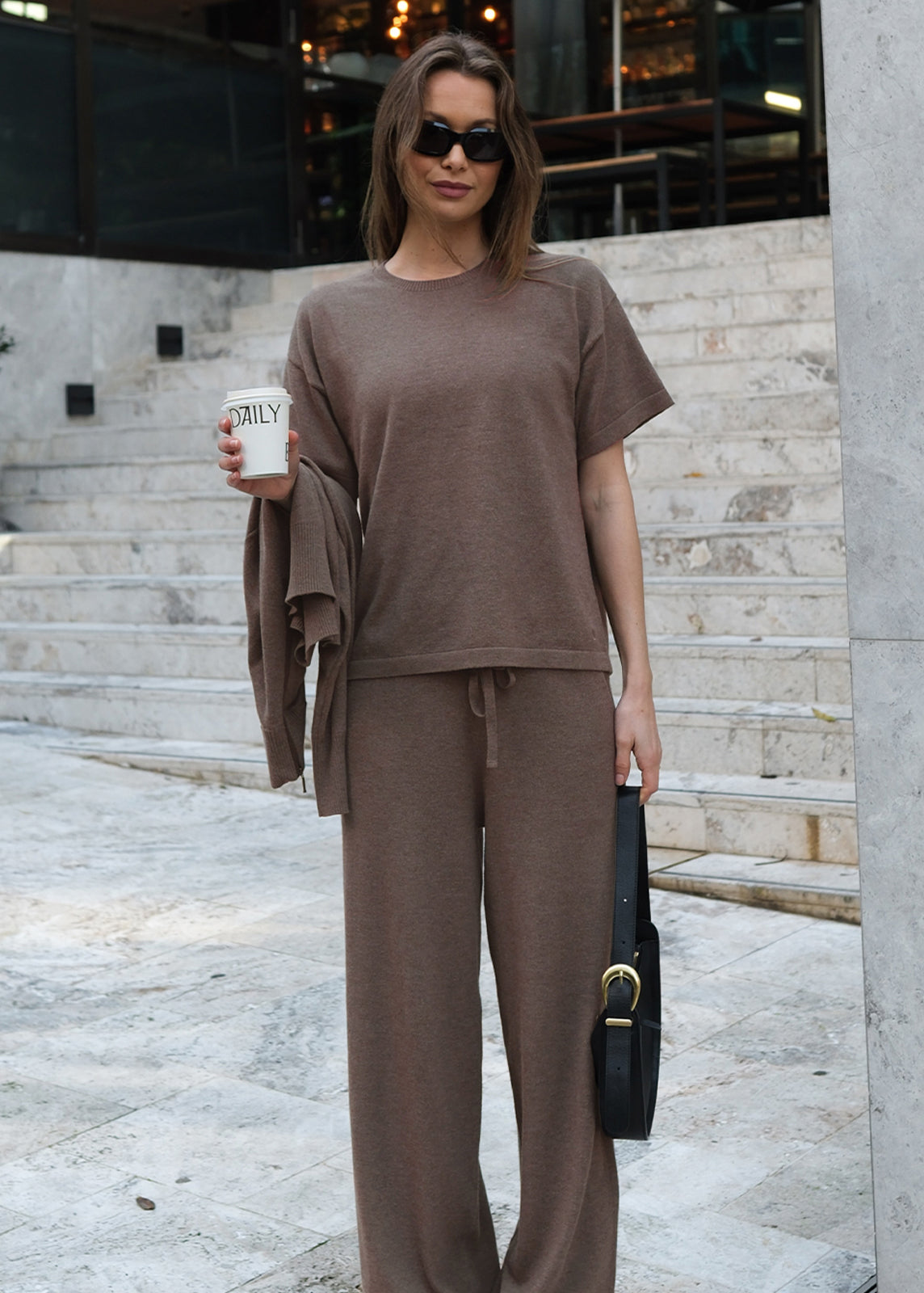 Model wearing brown knit lounge set with short-sleeve top, holding coffee cup and handbag
