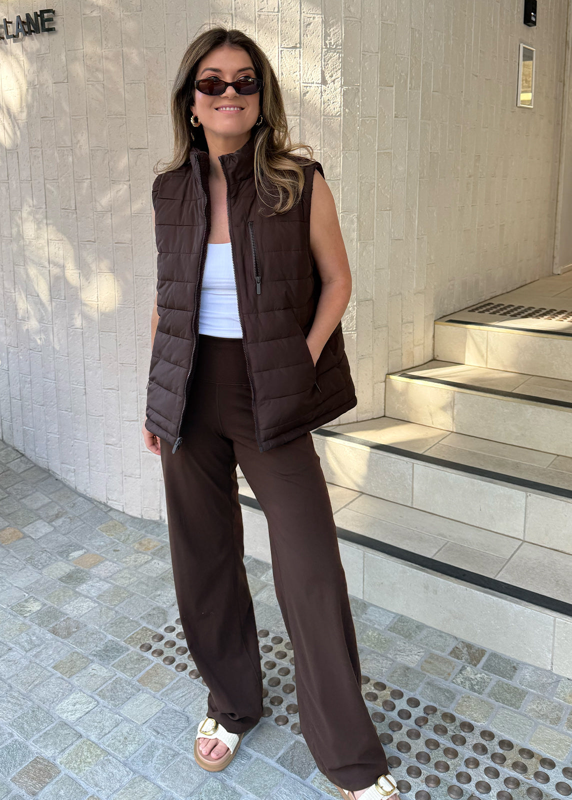 Woman wearing chocolate brown wide-leg pants and puffer vest, styled with slides for casual everyday wear