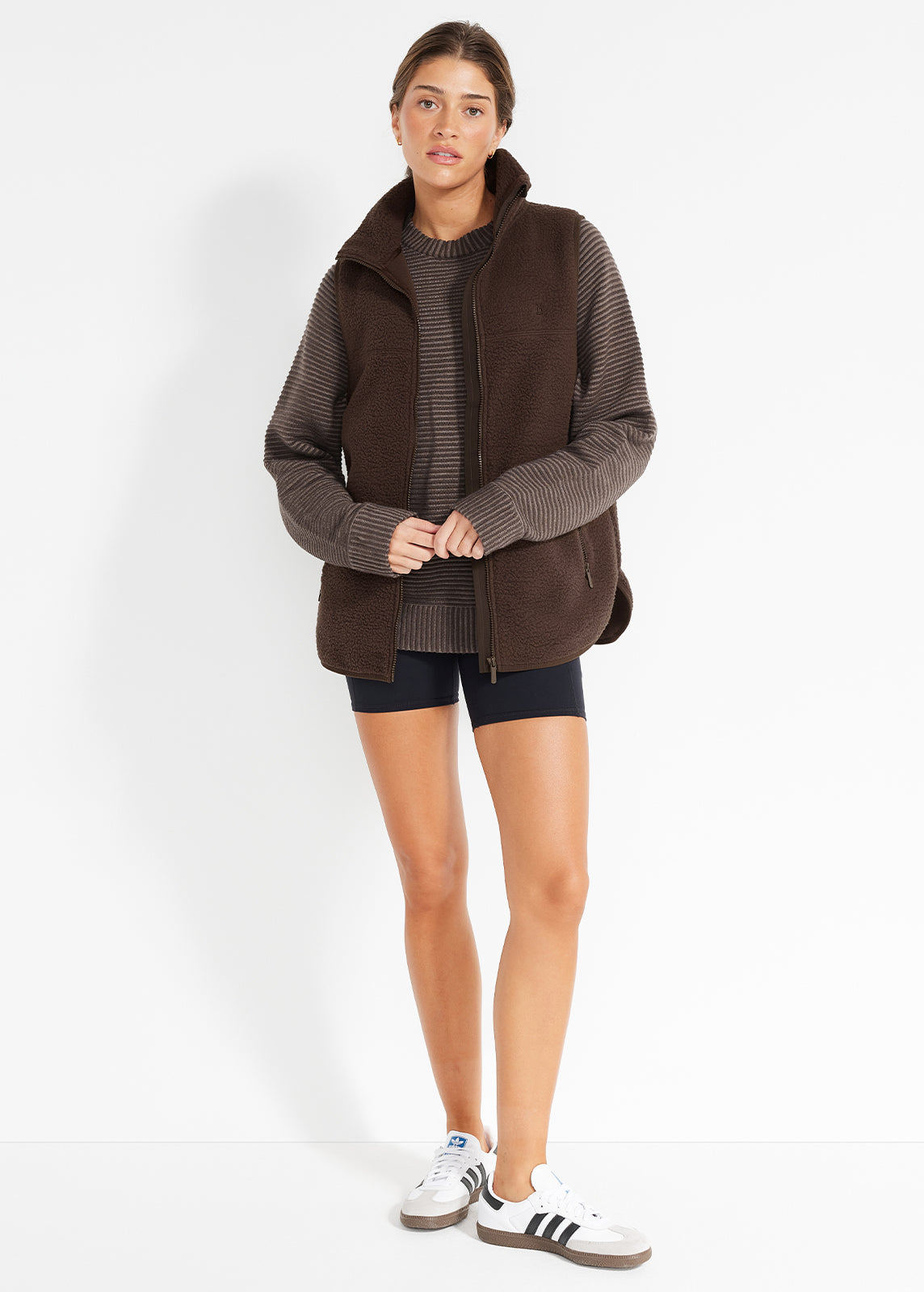 Model wearing brown ribbed knit jumper layered with fleece vest and black bike shorts