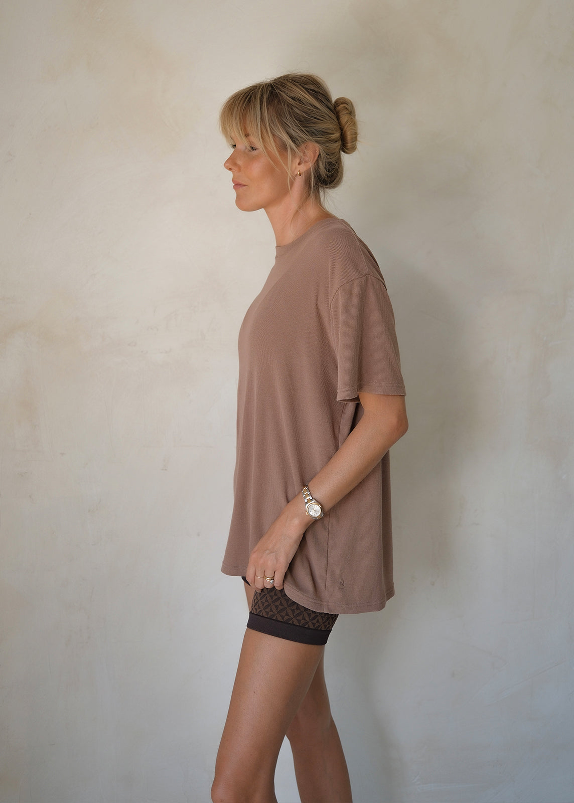Close Up Of Model Stood Facing Sideways Wearing A Walnut Coloured Oversized Tee With Crew Neckline, Short Sleeves And All-Over Ribbed Detail.