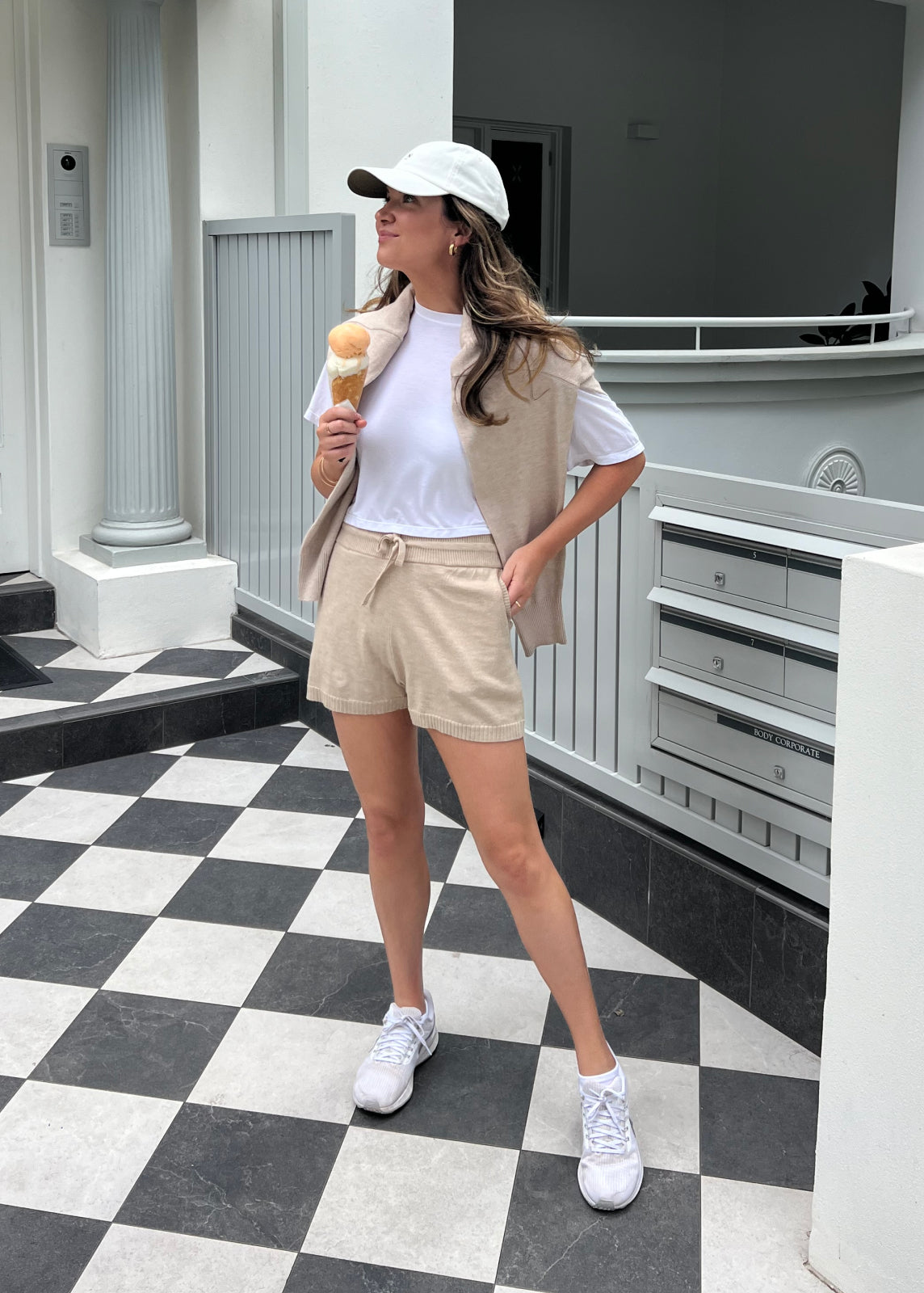 Model Stood Facing Sideways Wearing Oatmeal Coloured Knit Shorts With Pockets At The Hip And Drawstring To The Waist With A Matching Long Sleeve Knit Over Shoulders And A White Short Sleeve Tee.