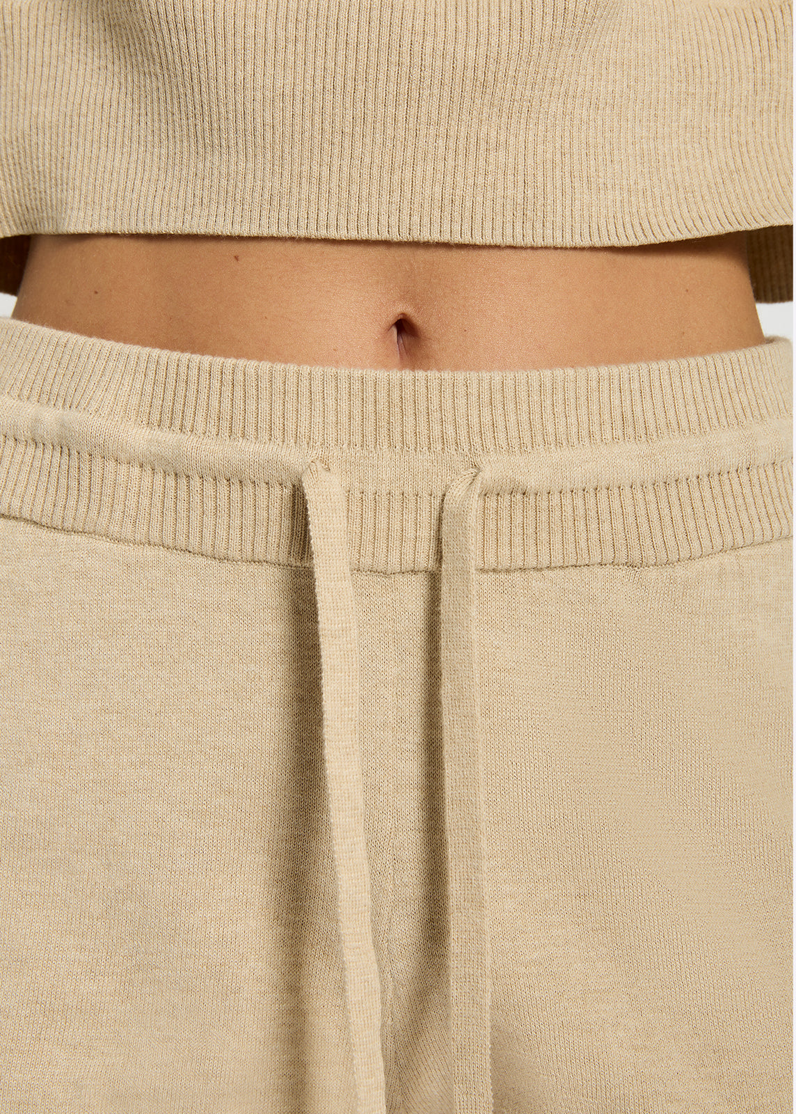 Close up of model wearing oatmeal coloured knit shorts with mid-thigh length, pockets showing the waistband with drawstring detail.