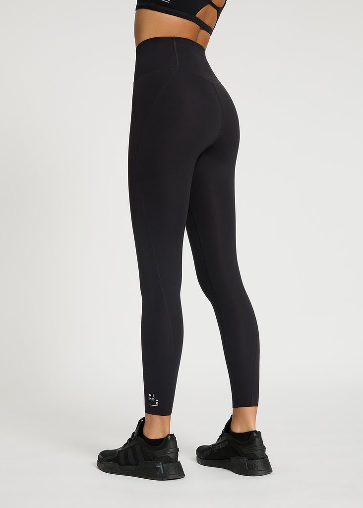 Close up of model stood facing backwards wearing black high rise leggings with hidden pocket detail to the back, 7/8 leg length and white logo detail to the bottom of leg